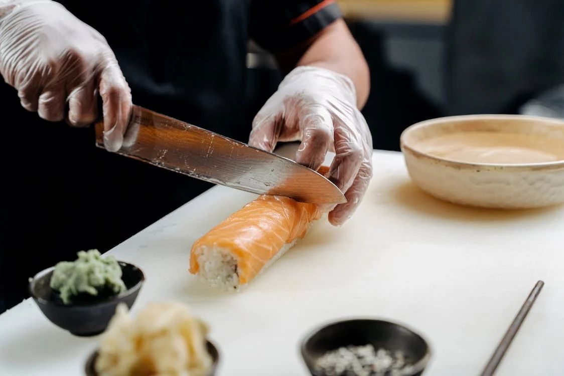 A chef in gloves slicing a salmon sushi roll with a sharp knife on a white cutting board, with wasabi, ginger, and soy sauce nearby.